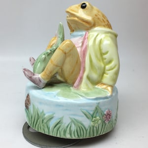 Peut inclure: Figurine de grenouille en céramique portant un manteau jaune, assise sur une base de nénuphar. La grenouille est brun clair avec des yeux noirs et tient un objet vert. La base est bleu clair avec de l'herbe verte et des fleurs roses.