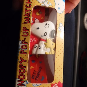 Puede incluir: Una caja amarilla y roja con un reloj Snoopy pop-up blanco en su interior. La caja tiene puntos blancos y las palabras "Snoopy Pop-Up Watch" en negro. El reloj presenta a Snoopy y Woodstock. La caja también dice "Best Friends!" y "Bonus".