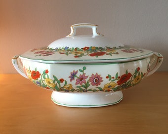 May include: A white porcelain serving dish with a lid, featuring a floral pattern in shades of red, yellow, pink, and green. The dish has a scalloped edge and two handles.