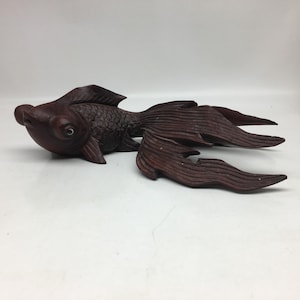 Op de afbeelding: Een donkerbruine, gesneden houten vissculptuur met gedetailleerde vinnen en schubben. De vis heeft een afgerond lichaam en een lange, vloeiende staart. De sculptuur ligt op een wit oppervlak.