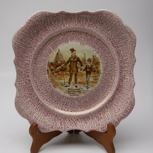 Puede incluir: Un plato de cerámica rosa con borde festoneado y una imagen impresa de dos hombres caminando en una escena de ciudad. El texto en el plato dice "Micawber Introduces David to London".