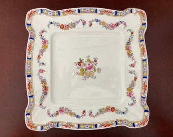 May include: A white square serving plate with a scalloped edge. The plate has a floral design in shades of pink, yellow, blue, and red. The border of the plate is decorated with a blue, yellow, and red pattern.