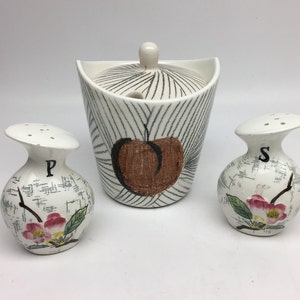 May include: A vintage ceramic salt and pepper shaker set with a matching sugar bowl. The shakers are white with black lettering that says "P" and "S" and feature a floral design. The sugar bowl is white with black and brown stripes and a brown apple design.