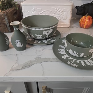 May include: A collection of sage green ceramic tableware with white relief designs. The set includes two small vases, two bowls, and two saucers. The bowls and saucers feature detailed scenes, while the vases have simpler relief patterns.