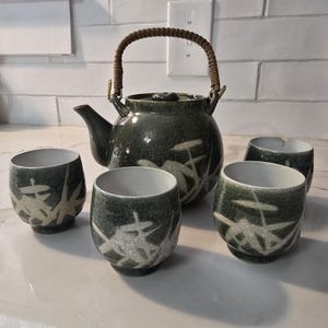 May include: A dark green ceramic tea set comprising a teapot and four cups. The teapot has a woven handle. The cups and teapot feature a white floral design. The set is displayed on a white countertop.