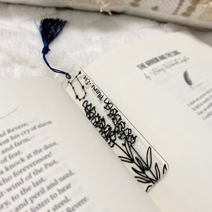 May include: A clear bookmark with a black floral design and the constellation Gemini. The bookmark has a blue tassel.