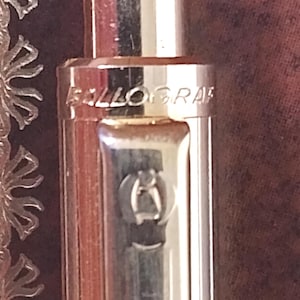 Puede incluir: Un bolígrafo de color oro rosa metálico con un mecanismo de botón pulsador cilíndrico en la parte superior. La palabra BALLOGRAF está grabada en el cuerpo superior y un pequeño emblema circular está grabado en el clip integrado del bolígrafo.