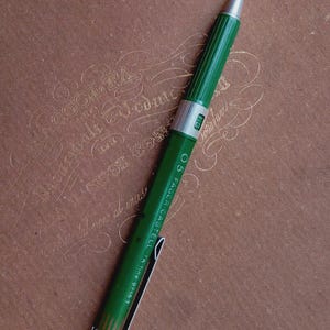 以下が含まれることがあります： 緑色のFaber-Castell TK-fine 9715 T製図用シャープペンシル。0.5の芯径表示があります。ペンシルにはリブ付きのグリップ部分、銀色の金属製ポケットクリップ、HBを表示する芯硬度表示窓、そして銀色のペン先が備わっています。