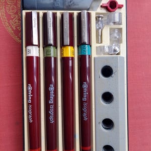 Puede incluir: Un juego de plumas técnicas Rotring Isograph en un estuche negro. Las plumas son de color rojo oscuro con detalles plateados y diferentes tamaños de punta, incluyendo 0,3, 0,35 y 0,70 mm. También se incluye tinta y accesorios.