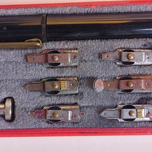 Könnte beinhalten: Ein Vintage-Kalligraphie-Stiftset in einem roten Etui. Das Set enthält einen schwarzen Stift, verschiedene Federn und andere Werkzeuge. Die Federn sind silberfarben und haben unterschiedliche Formen und Größen. Das Etui hat einen grauen Innenraum.