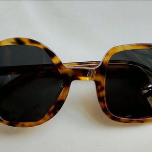 May include: Tortoiseshell-patterned sunglasses featuring an asymmetrical design with a circular left lens and a square right lens. The frames are made of a brown and black mottled acetate material with dark tinted lenses and small silver-tone rivets.