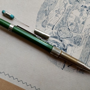 Op de afbeelding: Een groen vulpotlood met een zilveren metalen clip en een getextureerde metalen grip. Het potlood heeft een afneembare zilveren dop en een klein, los onderdeel met een blauwe punt en een dun draadje, liggend op een textieloppervlak.