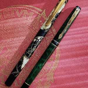 Könnte beinhalten: Zwei Füllfederhalter sind abgebildet. Ein Stift hat einen schwarzen Schaft mit einer detaillierten weißen Weltkarte und einem goldenen Clip. Der zweite Stift hat einen marmorierten dunkelgrünen Schaft mit einem goldenen pfeilförmigen Clip.