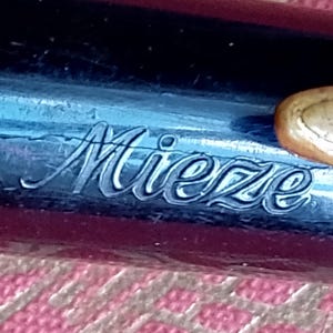 Op de afbeelding: Close-up van een pen met de naam "Mieze" gegraveerd in een cursief lettertype. De pen heeft een donkerblauwe en bordeauxrode behuizing met een goudkleurige accent in de buurt van de bovenkant. Het oppervlak van de pen vertoont tekenen van slijtage.