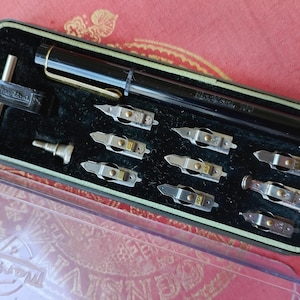 Puede incluir: Una pluma estilográfica negra con un clip dorado, acompañada de ocho plumines metálicos intercambiables y dos pequeños componentes metálicos, todo organizado en un estuche rectangular con interior de terciopelo negro y tapa de plástico transparente.