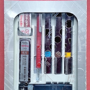 以下が含まれることがあります： 透明な窓が付いた白い箱に入ったRotring College製図セット。セットには、赤いシャープペンシル、色付きリングが付いた3本の黒い製図ペン、0.5 HBの芯ケース、消しゴムが含まれています。箱には定規が印刷されています。