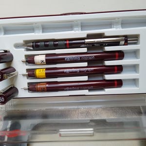 Könnte beinhalten: Ein Set aus Rotring Isograph technischen Stiften und einem Tikky 0,5 Druckbleistift in einem weißen und weinroten Kunststoffetui. Die Stifte haben unterschiedliche Strichstärken, die durch Zahlen auf den Kappen und Schäften angegeben werden.