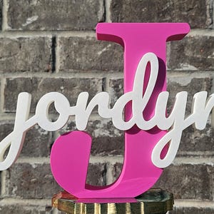 May include: A vibrant pink wooden letter "J" with the name "Jordyn" in white script. The personalized name is layered over the letter, creating a 3D effect. The sign is displayed against a brick wall.
