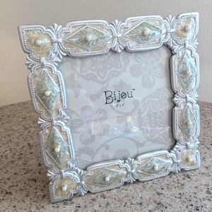 May include: A square picture frame with a silver-toned border featuring ornate scrollwork and diamond-shaped insets in cream and sage green. Each diamond contains a small white bead. The center insert displays the text Bijou 4 x 4.
