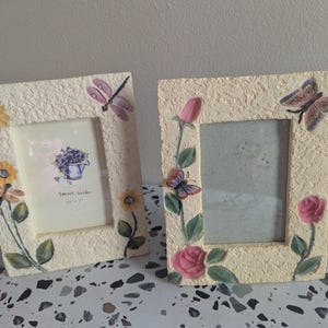 May include: Two decorative cream-colored picture frames. One frame features sunflowers and a dragonfly, the other has roses and butterflies. The frames have a textured finish and are designed to hold a 5" x 7" photo. The text "Secret Garden" is visible on one frame.
