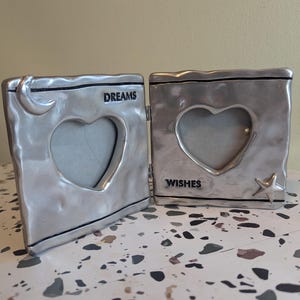 May include: Silver-toned metal picture frame with two heart-shaped openings. The frame is hinged and opens to reveal two separate picture frames. The left frame has a crescent moon design and the words "DREAMS" inscribed. The right frame has a star design and the words "WISHES" inscribed.