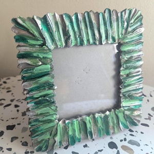 May include: Square picture frame with a textured, layered design. The frame is painted with shades of green and silver, creating a unique, artistic look. The frame is designed to hold a square photo.