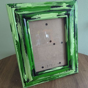 May include: A green picture frame with black and white paint accents. The frame is made of wood and has a distressed look. The frame is sitting on a wooden surface.