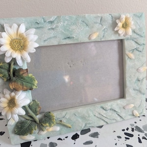 May include: A rectangular photo frame with a light blue, textured surface. The frame is decorated with white and yellow daisy flowers and green leaves. Small, white petal accents are also present.
