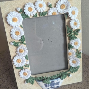 May include: A rectangular picture frame with a textured cream-colored border decorated with three-dimensional white daisy flowers, green leaves, and a blue and white patterned planter at the bottom. The frame features a central rectangular opening.