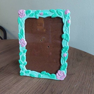 May include: A light green picture frame with a floral design. The frame is decorated with pink roses and green leaves.
