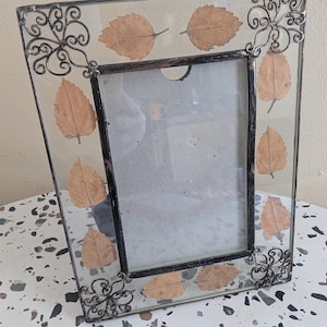 May include: A rectangular glass photo frame with a dark metal trim and decorative corner accents. The frame features dried leaves embedded within the glass. The frame is angled, showcasing its design and the neutral background.