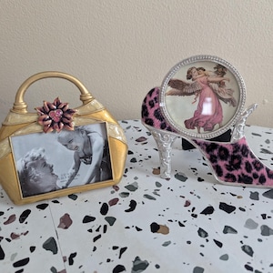 May include: Two decorative picture frames. One is a gold handbag shape with a pink flower accent. The other is a high-heeled shoe with a pink leopard print and a round frame with an angel image.
