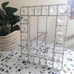 May include: A decorative silver metal photo frame with clear crystal accents. The frame has a rectangular shape and a wire grid design, with crystals along the edges. A small plant in a woven basket is in the background.