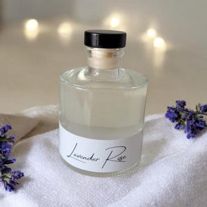 May include: A clear glass bottle with a black cap and a white label featuring the text Lavender Rose in black script. The bottle contains a pale liquid and is surrounded by small clusters of purple flowers on a white textured fabric surface.
