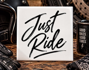 May include: A square white ceramic tile featuring the black handwritten-style text Just Ride centered on the front. The tile has a smooth, glossy finish and is positioned upright, displaying bold, dark lettering against a clean white background.