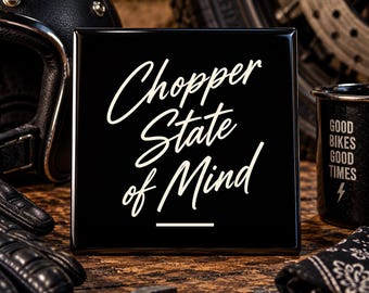 May include: A square black sign with white cursive text that reads Chopper State of Mind, featuring a horizontal line underneath the text. The sign is positioned among motorcycle gear including a black helmet, gloves, and a metal mug.