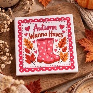 Op de afbeelding: Een vierkante witte keramische tegel met een rode hartjesrand en de tekst Autumn Wanna Haves in het rood. Het midden toont een paar roze regenlaarzen versierd met witte hartjes, omringd door oranje en rode herfstbladeren op een witte achtergrond.