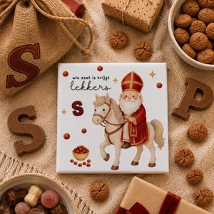 Op de afbeelding: Een vierkante witte tegel met een gekleurde illustratie van een figuur in een rood bisschopsgewaad en mijter op een wit paard. De tekst luidt "wie zoet is krijgt lekkers" met kleine bruine cirkels, sterren en een kom met snoepgoed.