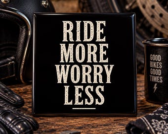 May include: A square black sign with white distressed lettering that reads RIDE MORE WORRY LESS, featuring a horizontal line underneath the text. The sign has a glossy finish and is positioned in a setting with motorcycle-related items.