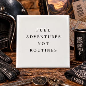 May include: A square white ceramic tile featuring the black printed text FUEL ADVENTURES NOT ROUTINES centered on the surface. The tile has a smooth, glossy finish and clean edges, presenting a clear and legible message in a simple serif font.