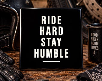 May include: A square black sign with white text that reads RIDE HARD STAY HUMBLE. The sign features a horizontal line below the text. The lettering is in a clean, sans-serif font, and the surface of the sign has a smooth, glossy finish.