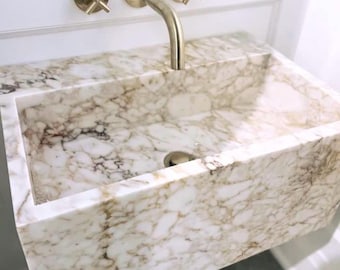 May include: A rectangular marble sink with a gold faucet and fixtures. The sink has a white and brown veined pattern. The faucet is curved, and the fixtures are cross-shaped. The sink is mounted on a white wall.