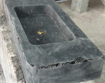 Puede incluir: Un lavabo rectangular de piedra de color gris oscuro con un borde áspero y sin terminar. El lavabo tiene un solo agujero de drenaje en el centro.