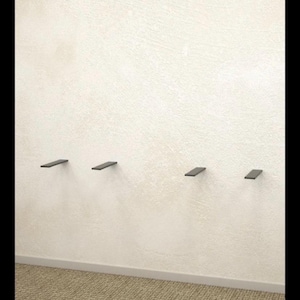 May include: Four small, rectangular, dark gray floating shelves are mounted on a textured, off-white wall. The shelves are evenly spaced and appear to be made of metal or a similar material. The bottom of the wall meets a beige carpet.