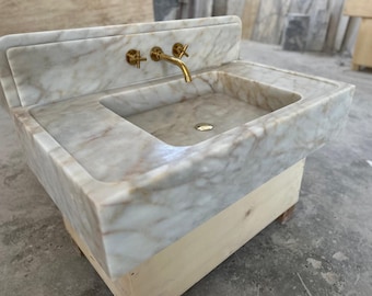 May include: A marble sink with a rectangular basin and a raised backsplash. The sink features a gold-colored faucet and fixtures. The marble has a white and tan veined pattern. The sink is mounted on a wooden base.