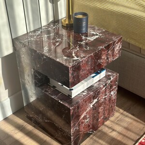 May include: A two-tiered red marble end table with a gold lamp and a black candle on the top tier. The bottom tier has a book titled "Simplicity" on it.