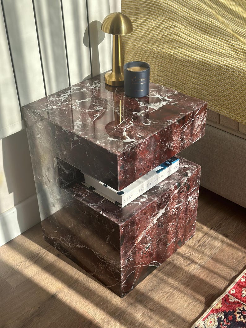 May include: A two-tiered red marble end table with a gold lamp and a black candle on the top tier. The bottom tier has a book titled "Simplicity" on it.