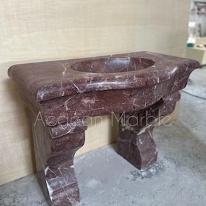 May include: A red marble bathroom vanity with a curved top and a sink. The vanity has two legs with ornate details.