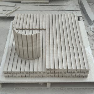 May include: A collection of light gray stone architectural elements, including rectangular and curved pieces, arranged on a wooden pallet. The pieces have a grooved, vertical design, suggesting a potential construction or design project. The overall aesthetic is minimalist and industrial.