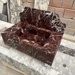 May include: A rectangular, red marble sink with white veining. The sink has a built-in backsplash and a grooved exterior design. The sink is made of stone and is a bathroom fixture.